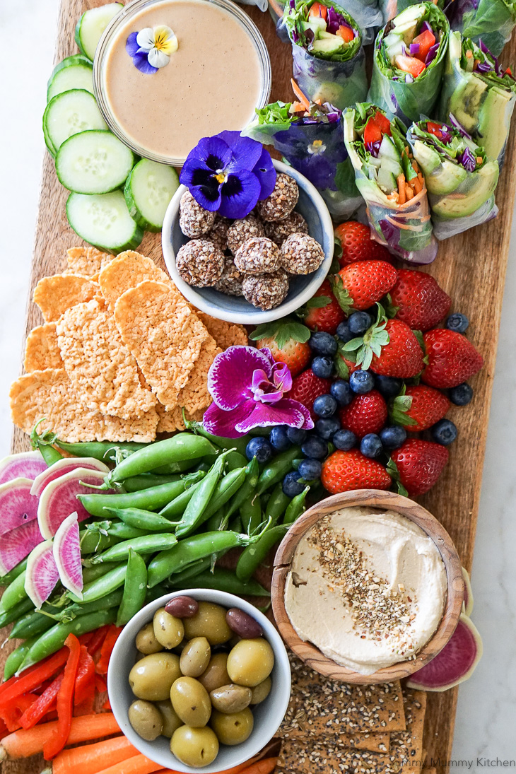 A beautiful vegetarian party platter snack board with fresh homemade vegan spring rolls, hummus and vegetables, olives, crackers, berries, and truffles. 