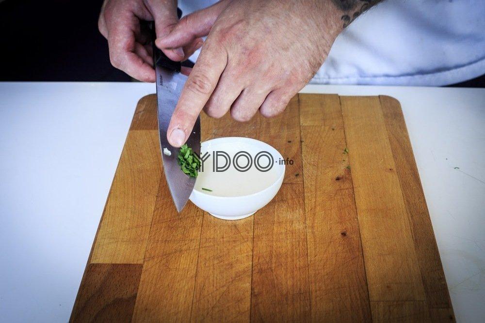 сметана в маленькой пиале на деревянной доске на столе, рядом с пиалой большой кухонный нож, на кончике ножа мелко нарезанная зелень