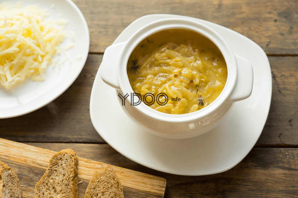 луковый суп в супнице на белой тарелке, рядом стоит тарелка с тертым сыром, кусочки багета лежат на деревянной доске