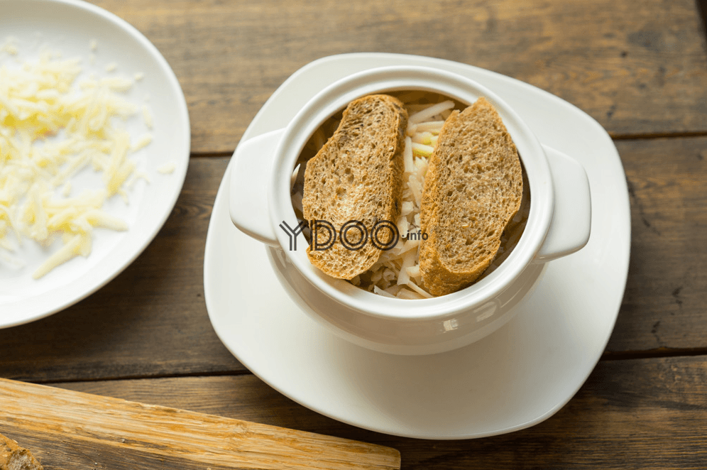 супница, наполненная луковым супом, сверху суп присыпан сыром и лежат два кусочка багета