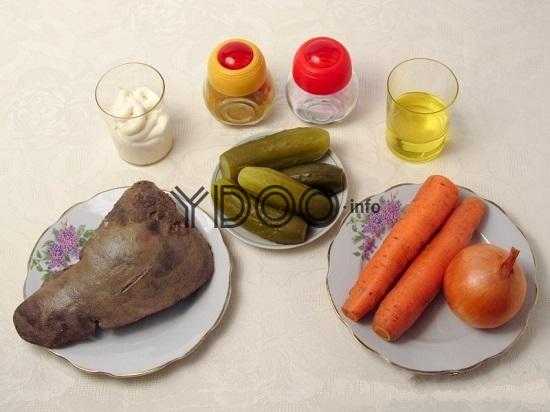 на столе стоит тарелка с говяжьей печенью, рядом на блюдце лежат соленые огурцы, на тарелке лежит две морковки и один репчатый лук в шелухе, в стакане растительное масло, рядом стоят две баночки со специями и стакан с майонезом