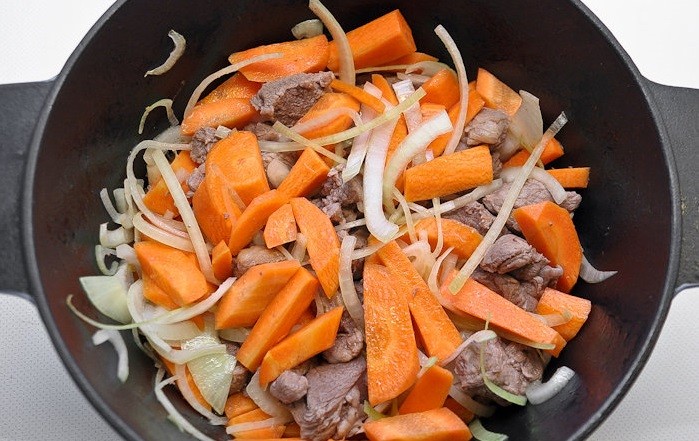 жареные куски мяса, перемешанные с нарезанным репчатым луком и морковью, в сковороде на столе