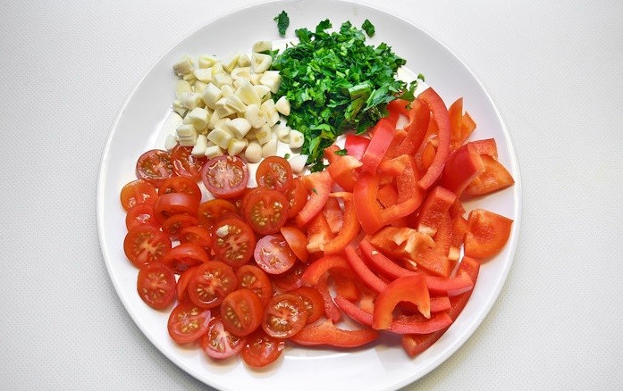 нарезанные помидоры черри, красный сладкий перец, зубчики чеснока и зелень на белой тарелке на столе