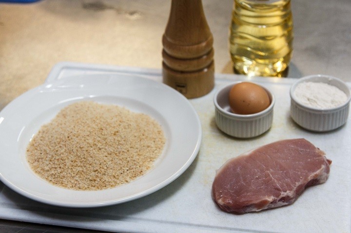 панировочные сухари на белой тарелке, кусок свинины, солонка, куриное яйцо в пиале на разделочной доске на столе, рядом бутылка растительного масла 