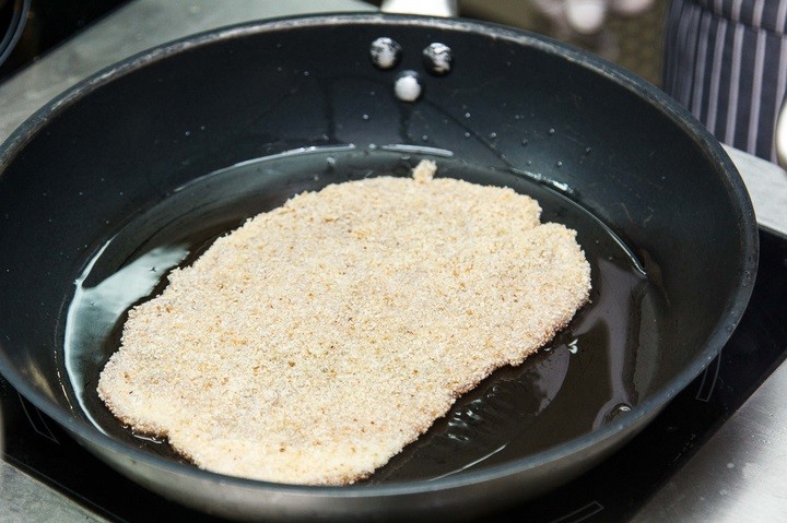 сырой кусок свинины в панировочных сухарях на сковороде с маслом на плите
